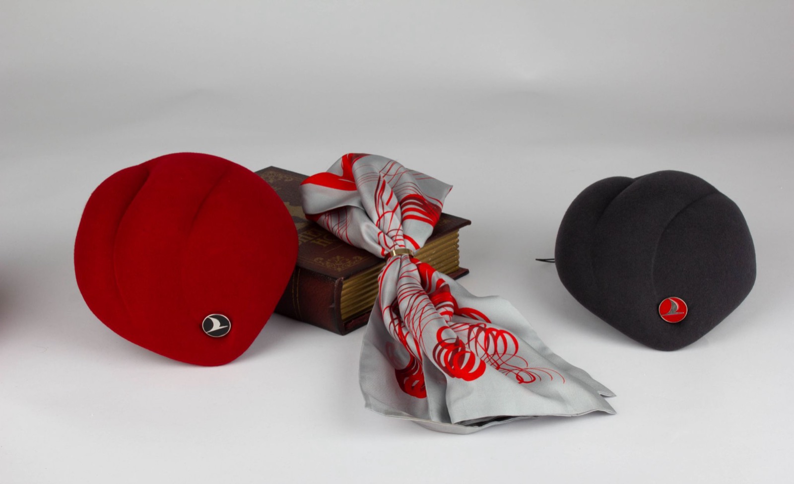 A red and a navy cabin-crew pillbox hat with an enamelled airline crest, beside a printed grey and red scarf