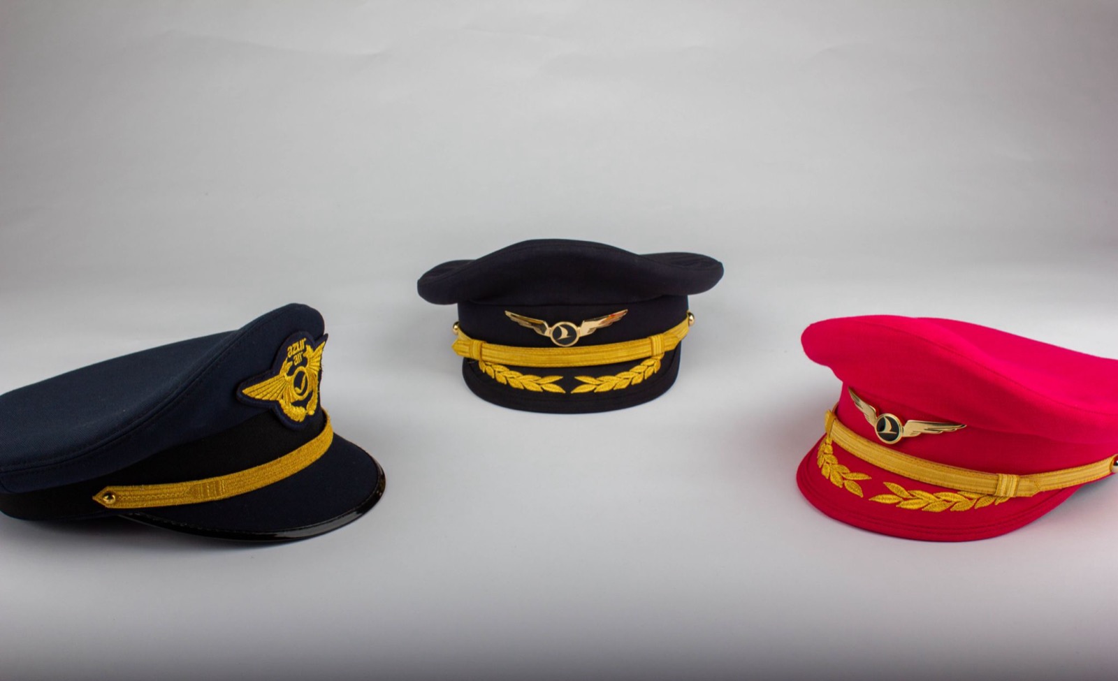 Three structured service caps, two navy and one pink, each with gold braid and a winged badge