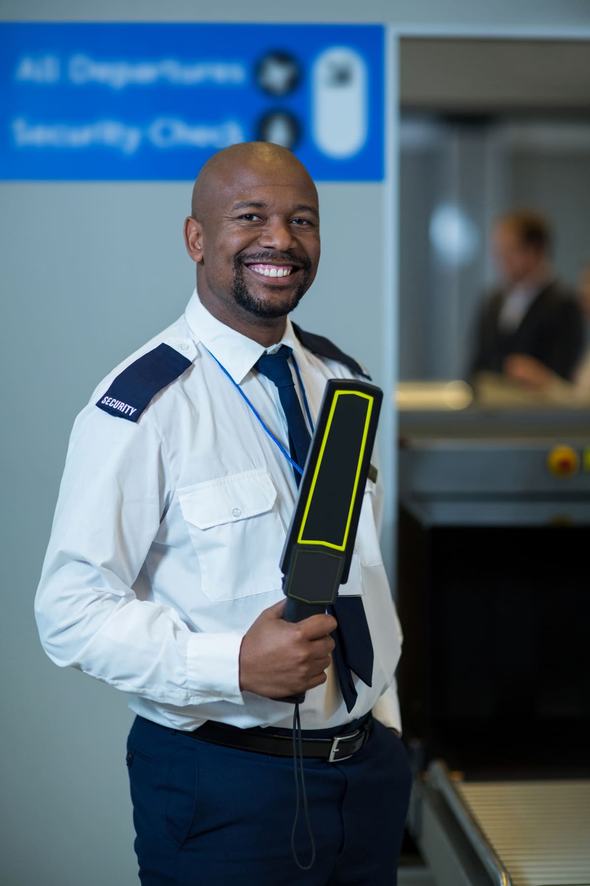 A smiling airport security officer holding a handheld metal detector at the checkpoint