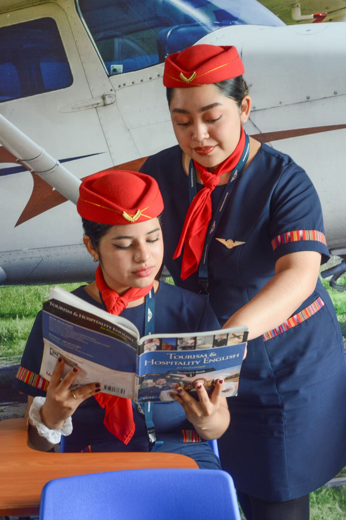 Two aviation students in navy uniforms with red caps studying together near a light aircraft